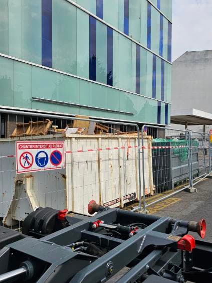 Benne remplie de bois de chantier derrière des barrières de sécurité au pied d'un immeuble