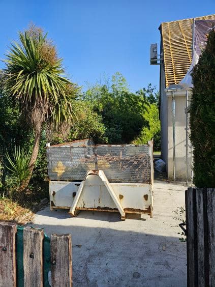 Benne positionnée dans une allée étroite entre une maison et un portail de jardin