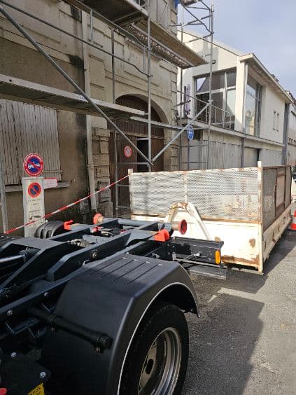 Camion porte-benne déposant une benne devant un immeuble en rénovation équipé d'un échafaudage